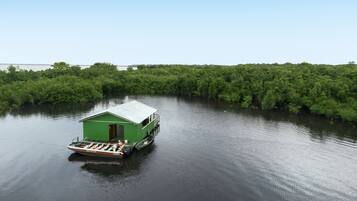 A green houseboat with a covered deck and a small cabin, floating on calm water surrounded by dense greenery.