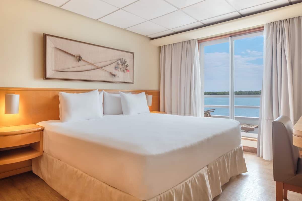 Habitación de hotel con una cama grande, cabecera de madera, mesita de noche con lámpara, vistas al agua a través de amplias ventanas y un cuadro en la pared.
