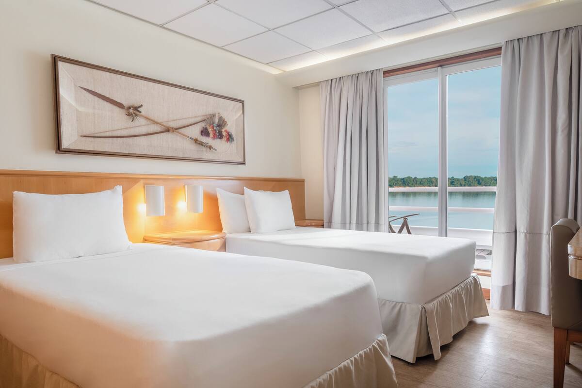 Habitación de hotel con dos camas, un ventanal con cortinas, un cuadro en la pared y vistas a un cuerpo de agua.