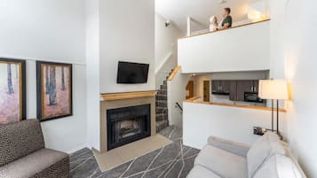 Een moderne woonkamer met een open haard, een bank, een aan de muur bevestigde televisie en een trap naar een hoger gelegen verdieping.