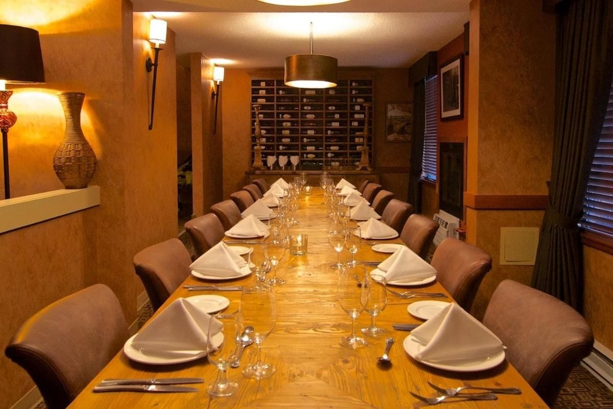 Ein langer Esstisch ist für eine Mahlzeit gedeckt, mit Stühlen an beiden Seiten, darüber ein Kronleuchter und im Hintergrund eine Weinwand.