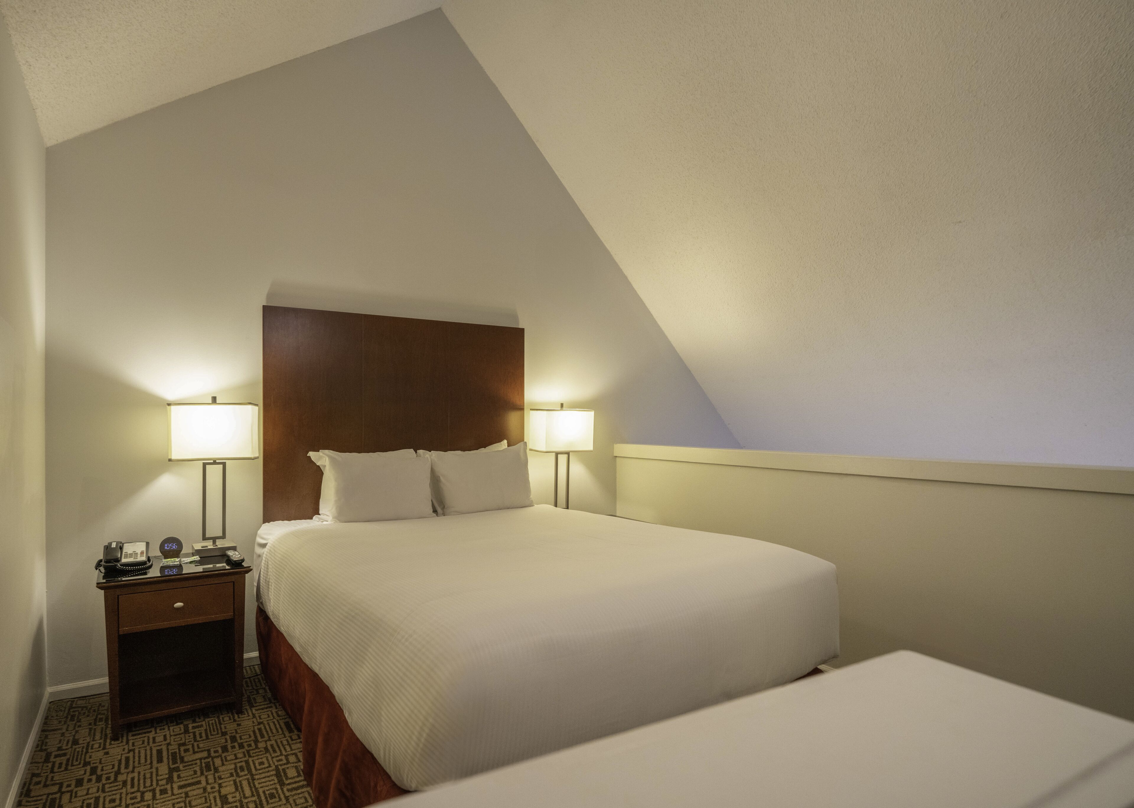 A hotel room with a large bed, two bedside lamps, a nightstand with a phone and a clock, and a patterned carpet.
