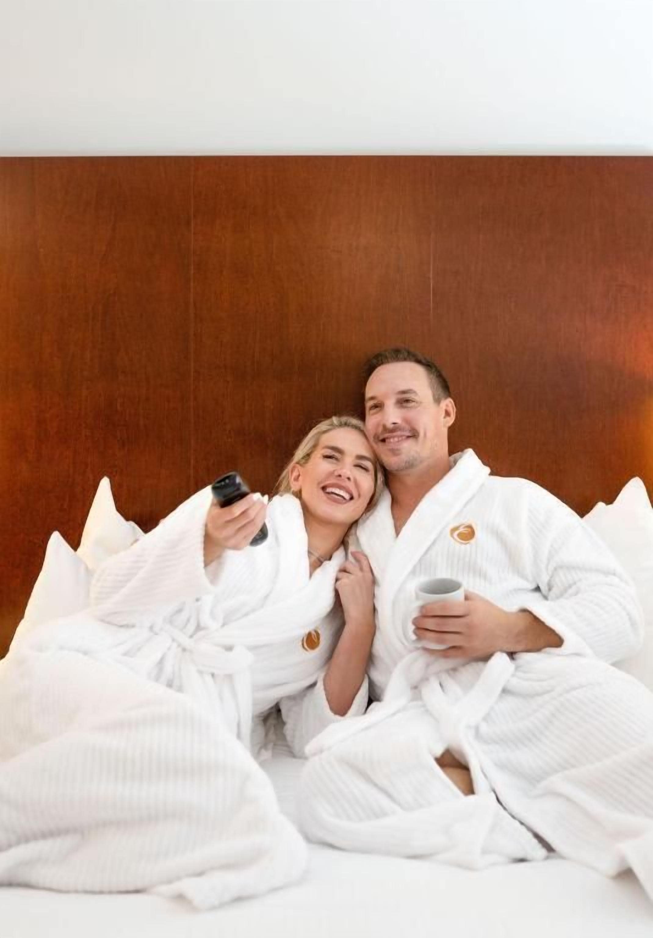 A couple in white bathrobes lying on a bed with a wooden headboard.