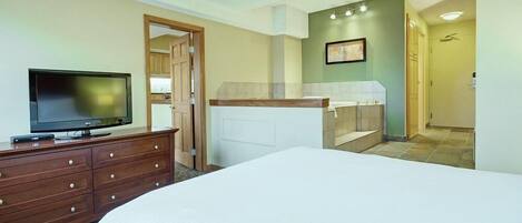 A hotel room with a bed, a TV on a dresser, a bathroom with a shower, and a door leading to another room.