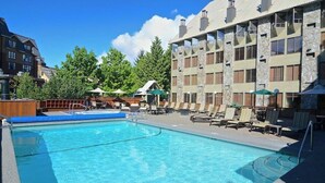 A swimming pool with lounge chairs and umbrellas, surrounded by a building with stone facade and multiple windows. - Mountain Side Hotel Whistler by Executive (Whistler)
