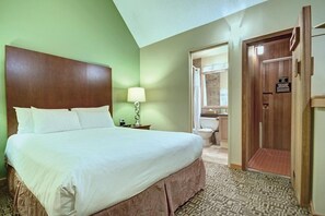 A hotel room with a large bed, a nightstand with a lamp, a bathroom with a shower, and a wooden door. - Mountain Side Hotel Whistler by Executive (Whistler)