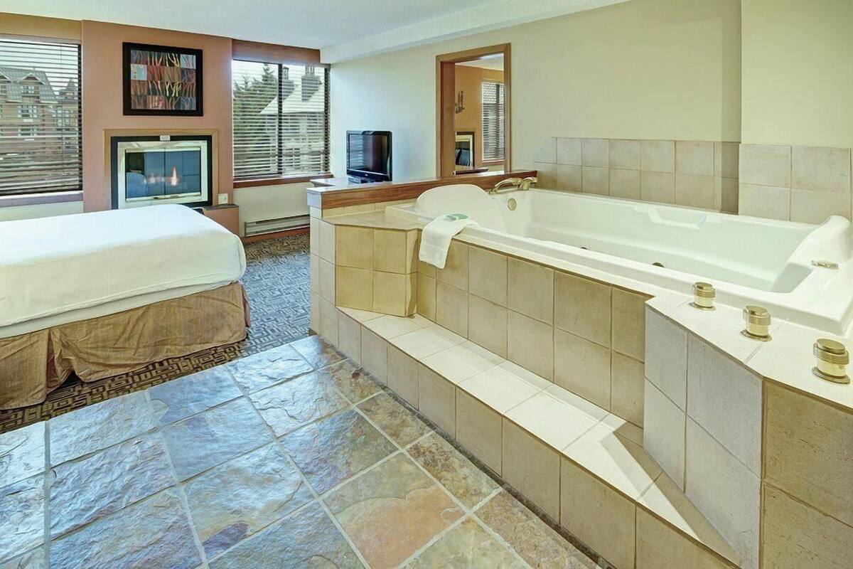 Habitación de hotel con una bañera grande, una cama, un televisor y una ventana con persianas.