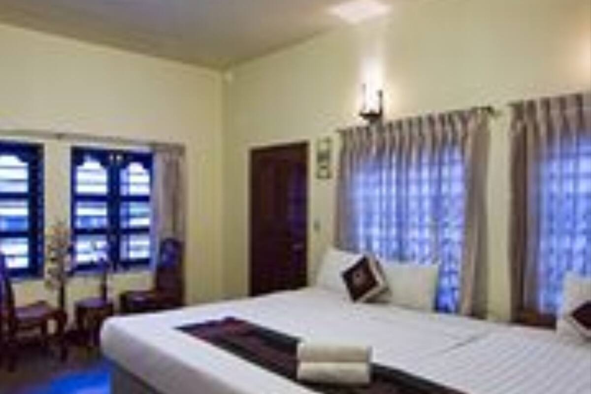 A hotel room with a large bed, a desk, and a chair. There are windows with curtains, a wall-mounted light fixture, and a door.