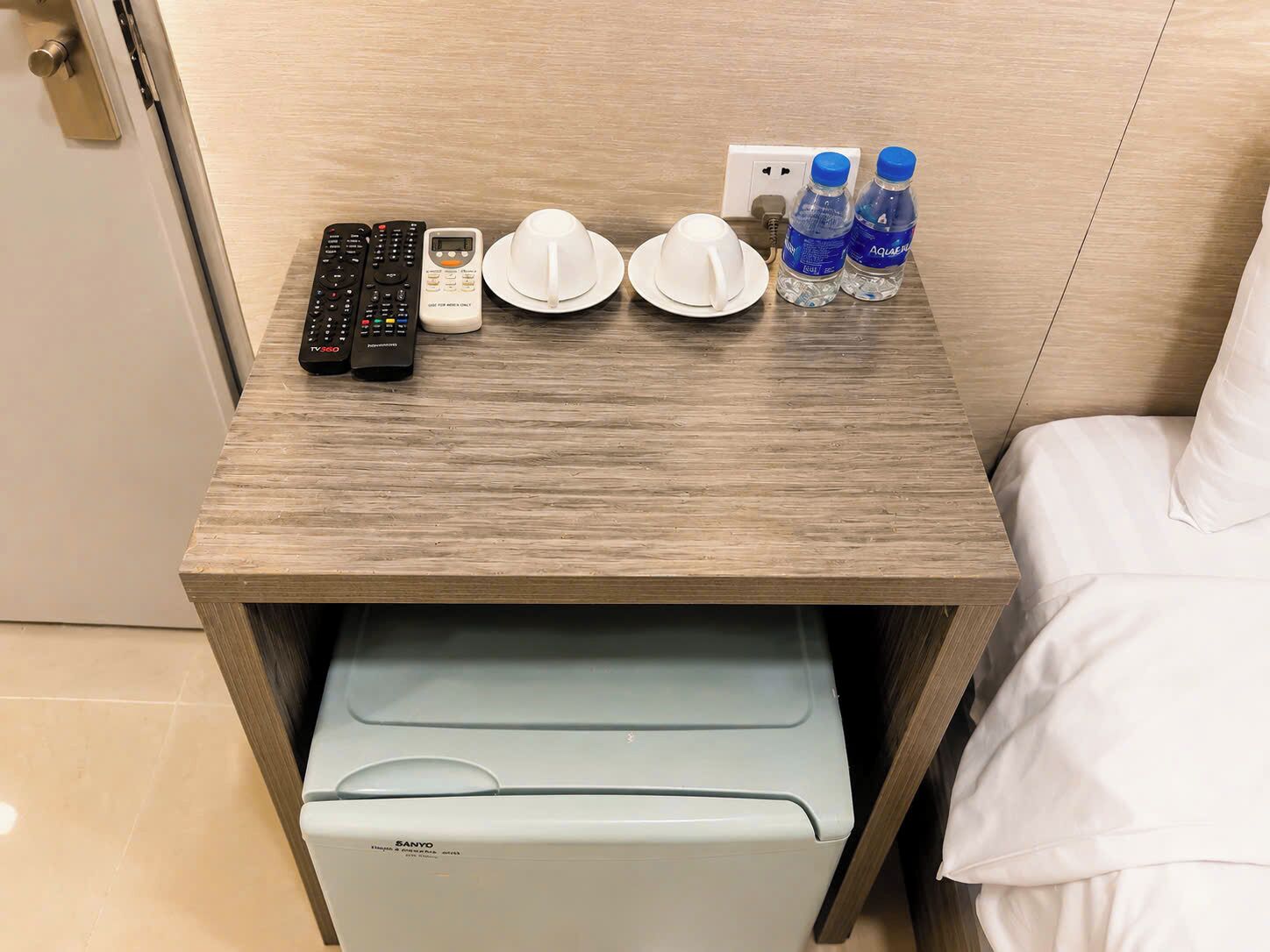 Habitación de hotel con una mesita de noche de madera, una impresora, dos mandos a distancia, dos gorras blancas y dos botellas de agua.