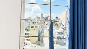 A window view of a cityscape with buildings, a clear sky, and a blue curtain partially covering the window. - Hikari Stay - Bui Vien CBD (Ho Chi Minh City)