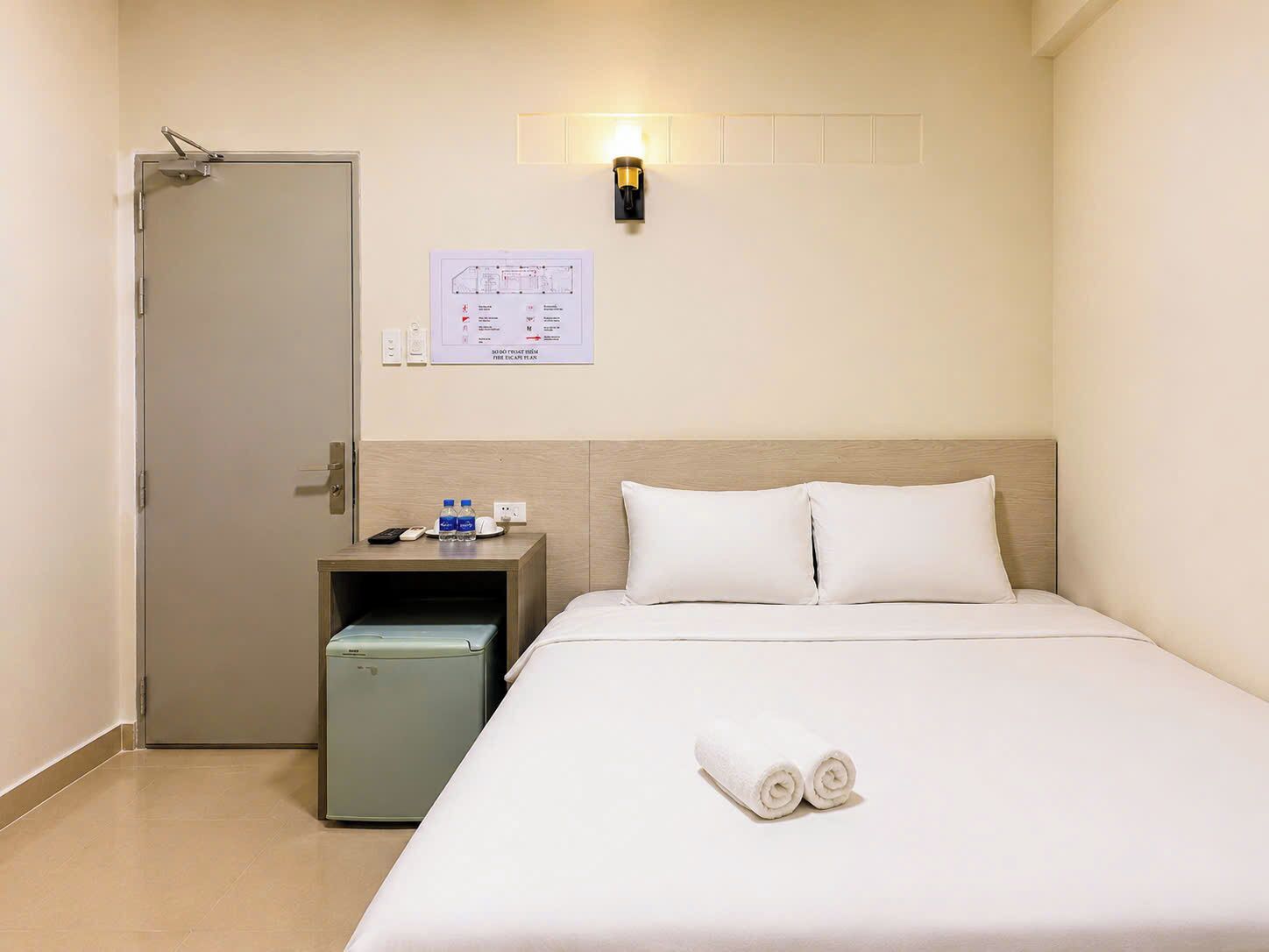 Une chambre d’hôtel avec un lit, une table de chevet, une lampe fixée au mur et une porte.