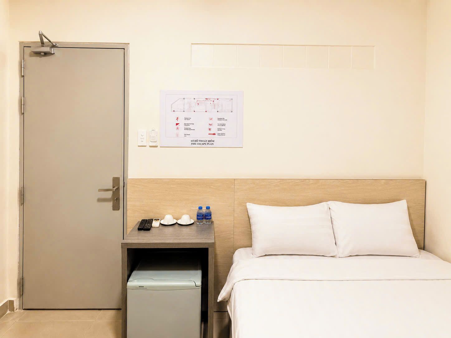 Ein Hotelzimmer mit einem Bett, einem Nachttisch mit Telefon, Fernbedienungen und Wasserflaschen; eine Tür mit Griff; ein an der Wand befestigter Fluchtplan; und ein kleines Gerät, möglicherweise eine Minikühlschrank.