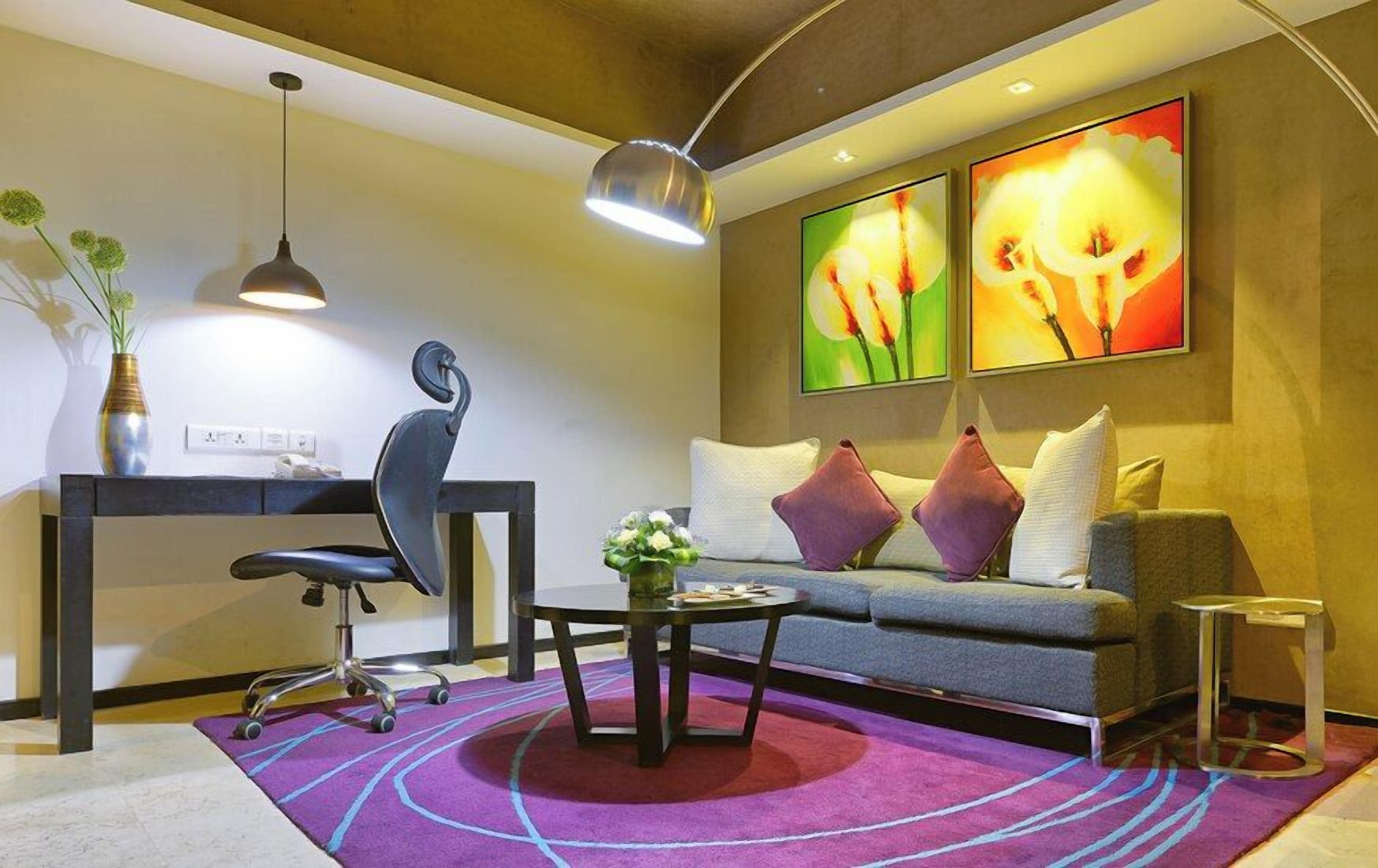 Un salon moderne comprenant un canapé, une table basse, un bureau et une chaise. Deux lampes sont fixées au mur, et un vase rempli de fleurs est posé sur le bureau.