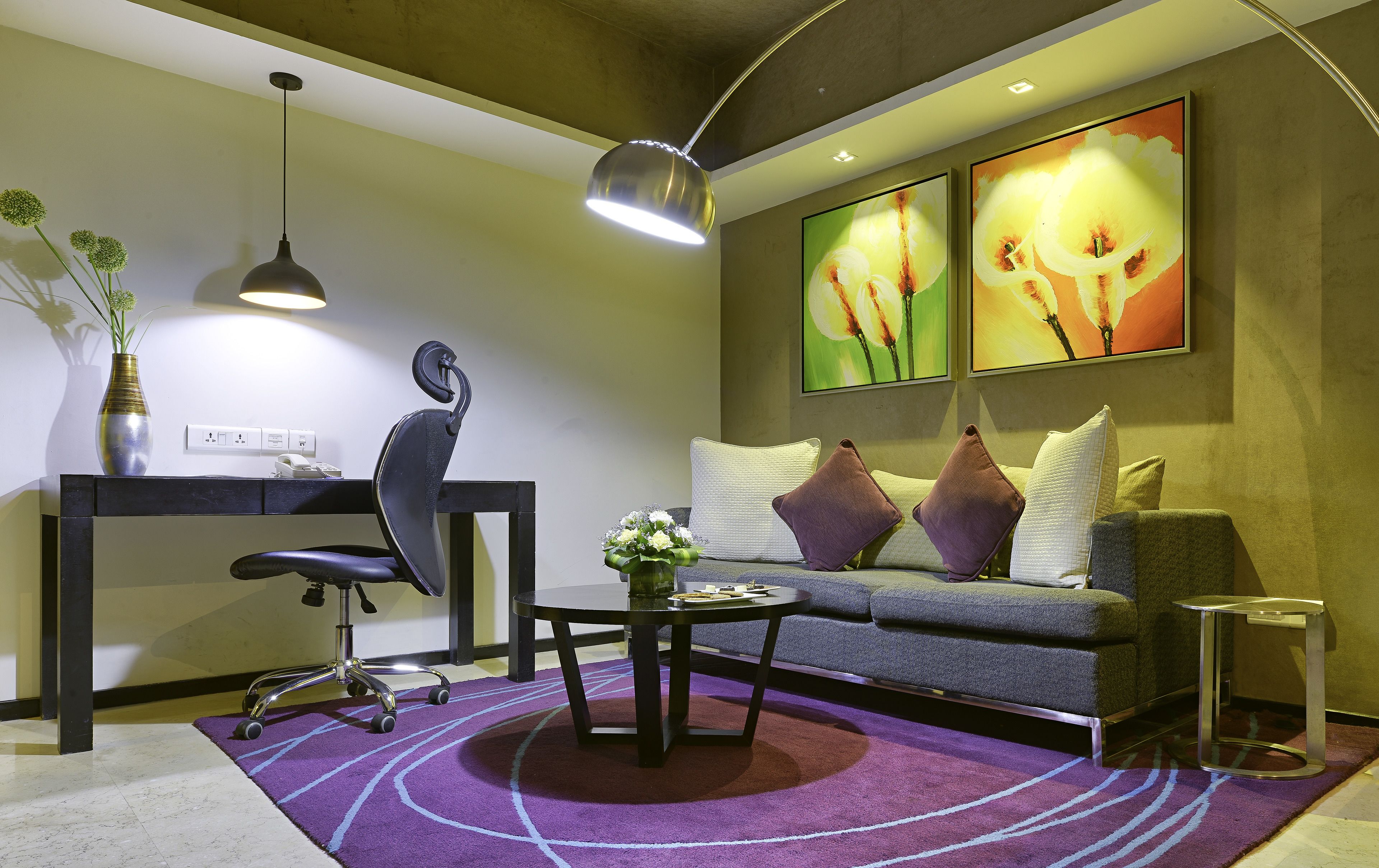 A modern living room with a sofa, coffee table, and a desk with a chair. There are two paintings on the wall and a vase with flowers on the desk.