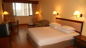 Une chambre d’hôtel avec un grand lit, deux lampes de chevet, une chaise, une petite table et une fenêtre avec des rideaux.