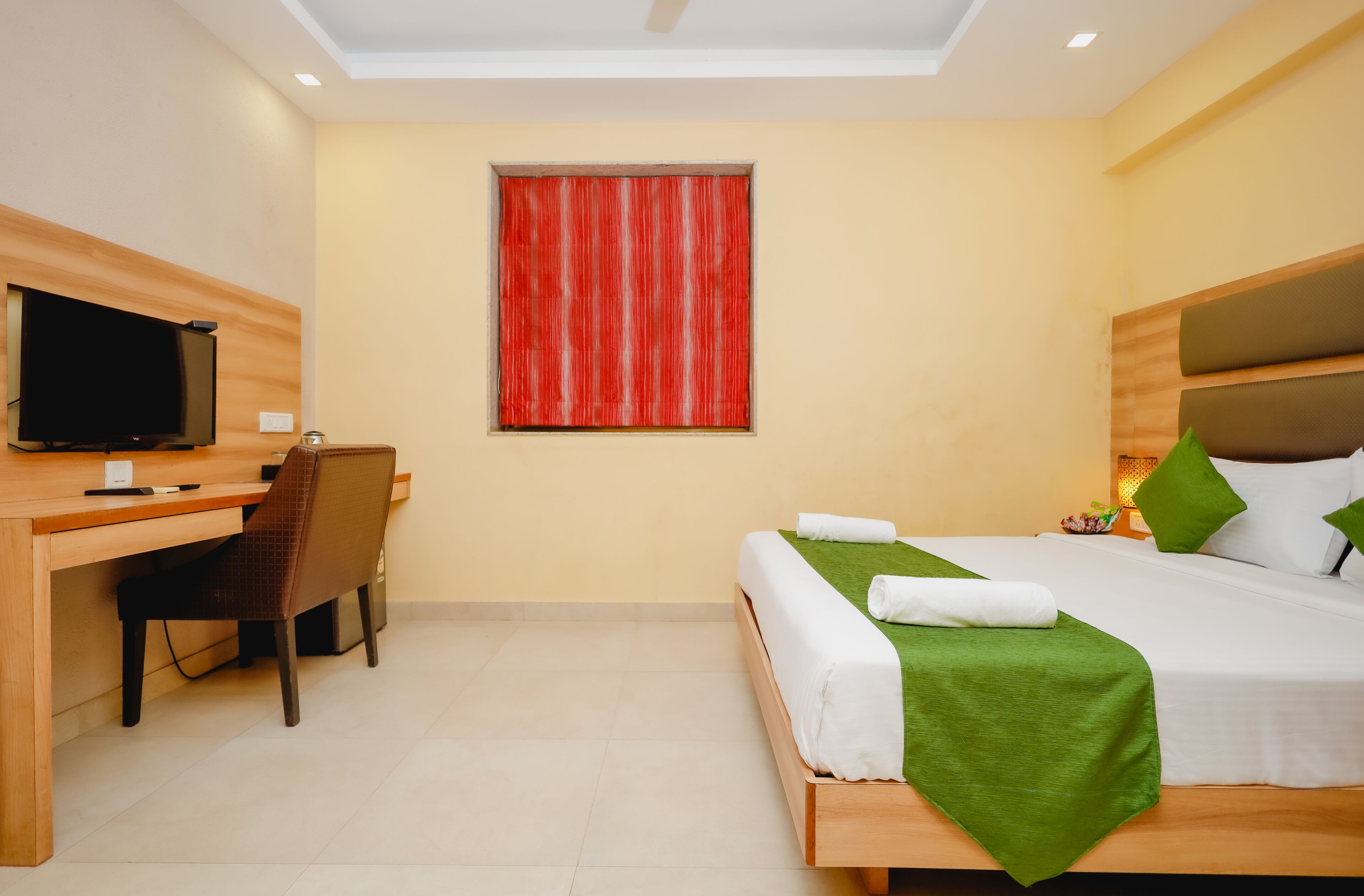 A hotel room with a bed, a desk, a chair, a television, and a window with red curtains.