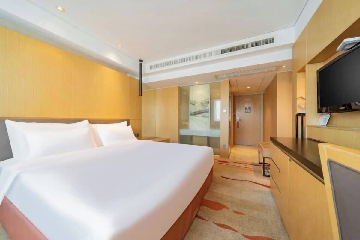 Ein Hotelzimmer mit einem großen Bett, einem Schreibtisch, einem Fernseher und einem durch eine Glaswand abgetrennten Badezimmer.