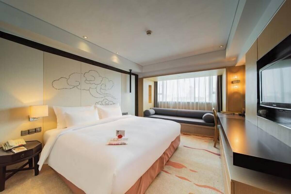 Ein Hotelzimmer mit einem großen Bett, einem Schreibtisch mit Lampe, einem Fernseher und einem Fenster mit Vorhängen.