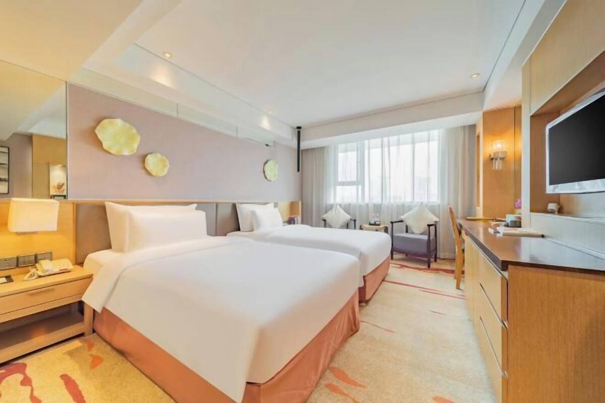 Ein Hotelzimmer mit zwei Betten, einem Schreibtisch, einem Fernseher und einem großen Fenster.