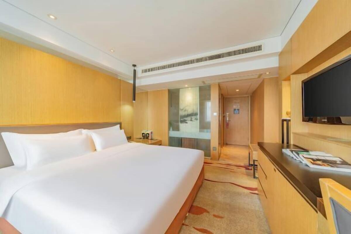 Ein Hotelzimmer mit einem großen Bett, einem Schreibtisch, einem Fernseher und Blick auf die Stadt durch eine Glastür.