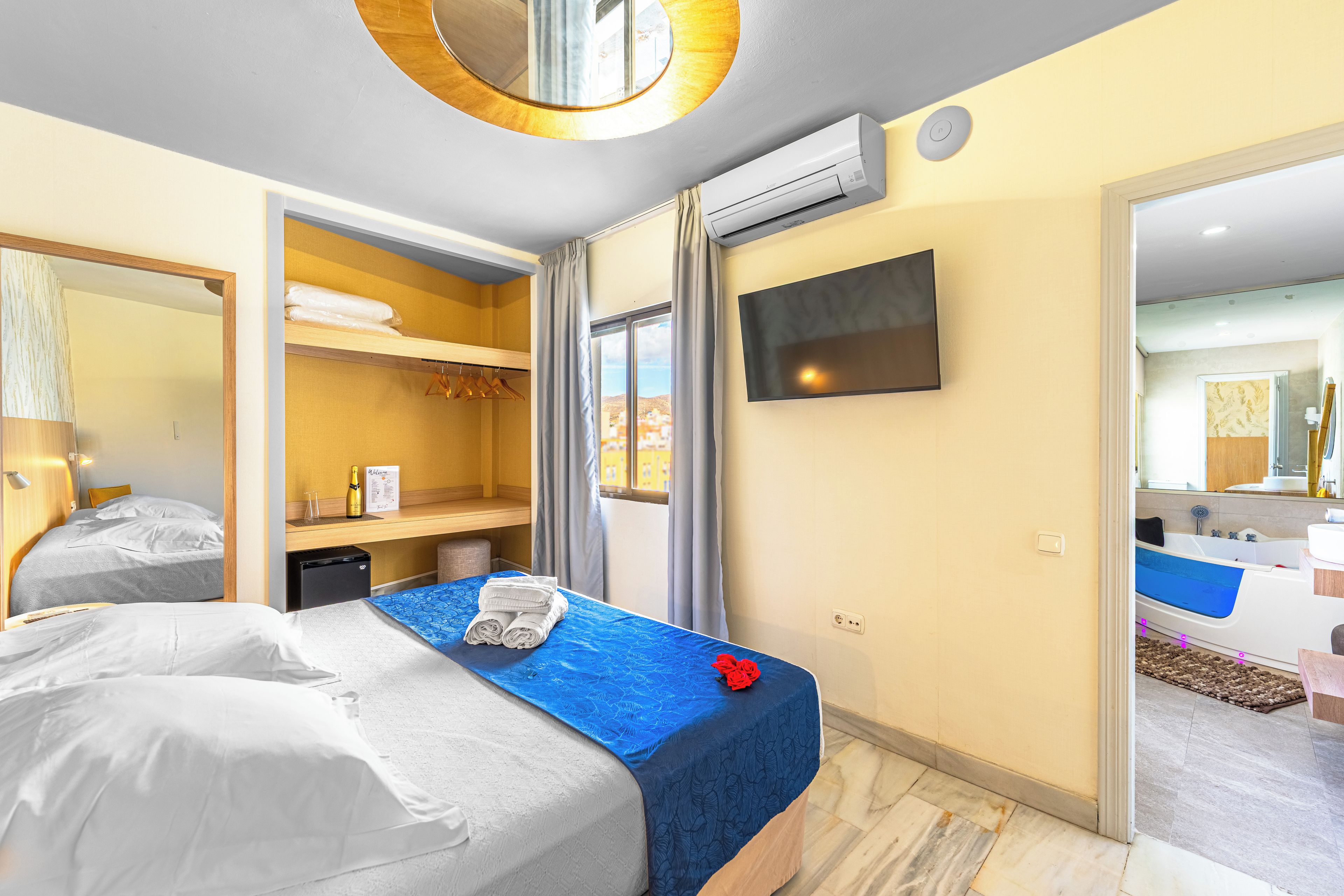 A hotel room with a bed, a desk, a wardrobe, a TV, and a bathroom visible through an open door.