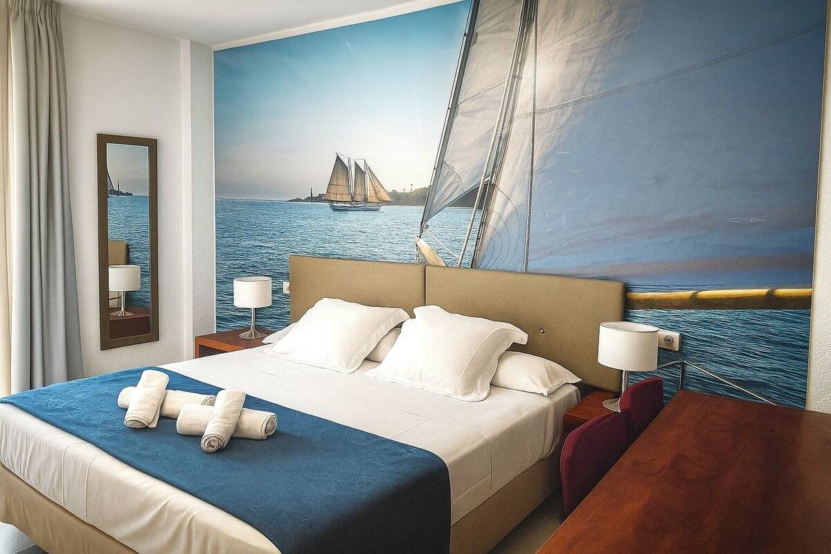 A hotel room with a large bed, a headboard with a sailboat painting, a nightstand with a lamp, and a view of the sea through the window.