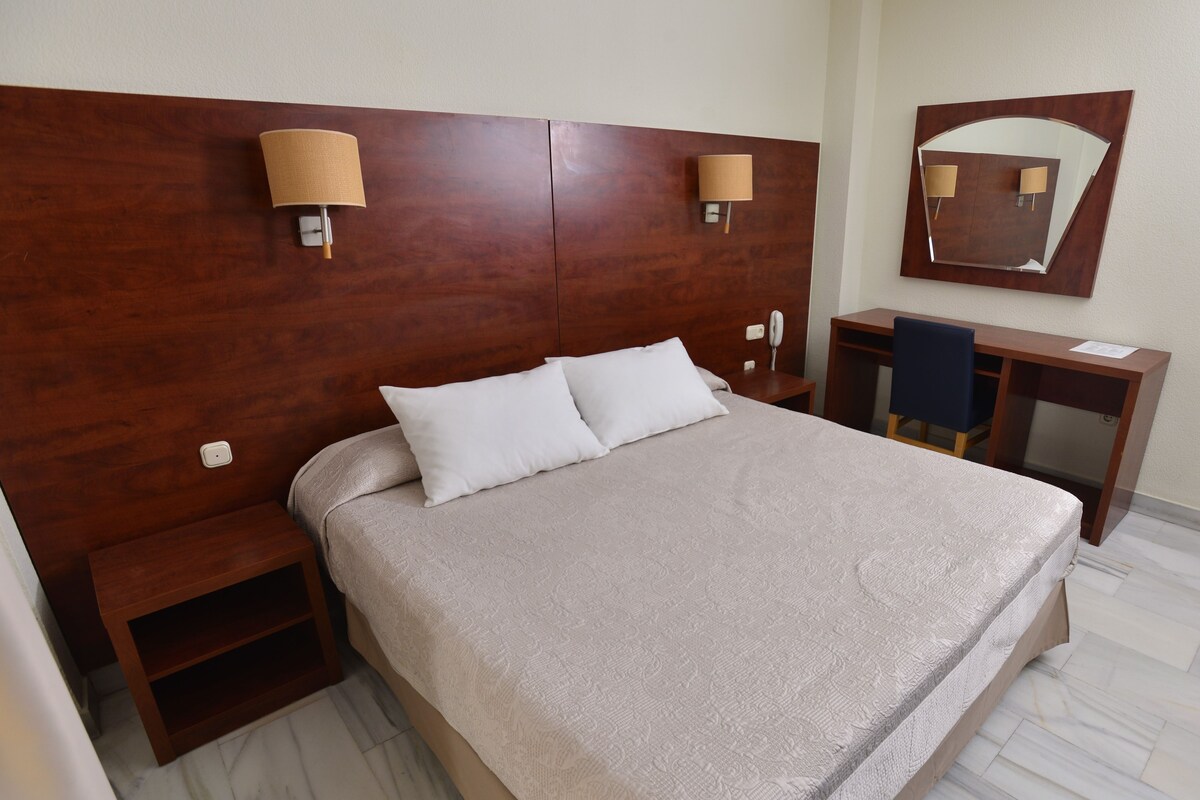 A hotel room with a large bed, two bedside tables, a desk, a chair, a mirror, and a telephone.