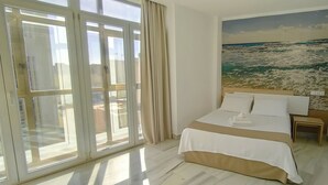Une chambre d’hôtel avec une grande fenêtre, un lit recouvert de draps blancs et un tableau représentant l’océan accroché au mur.