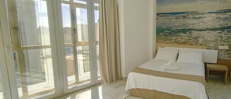 Un cuarto de hotel con un ventanal grande, una cama con ropa blanca y un cuadro del océano en la pared.