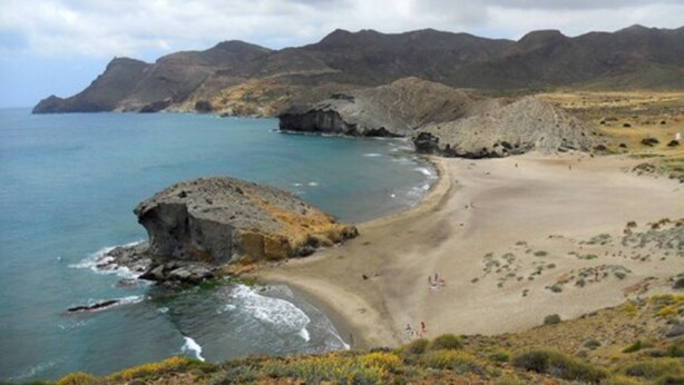 Un paysage côtier avec une plage de sable, des affleurements rocheux et des montagnes à l’horizon.