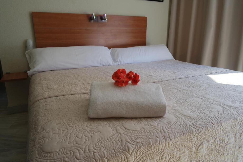 A neatly made bed with a floral-patterned bedspread, a folded towel, and a red flower decoration.