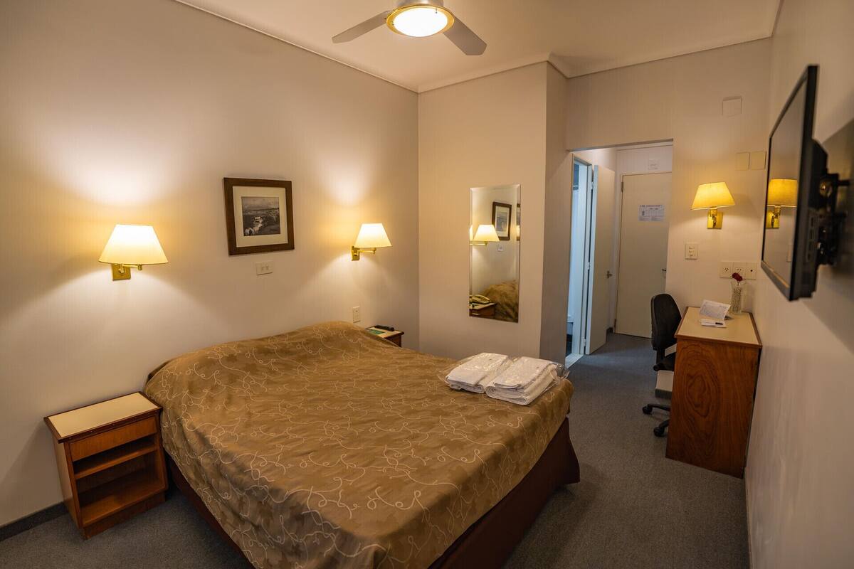 A hotel room with a bed, bedside lamps, a desk, a chair, a mirror, and a ceiling fan.