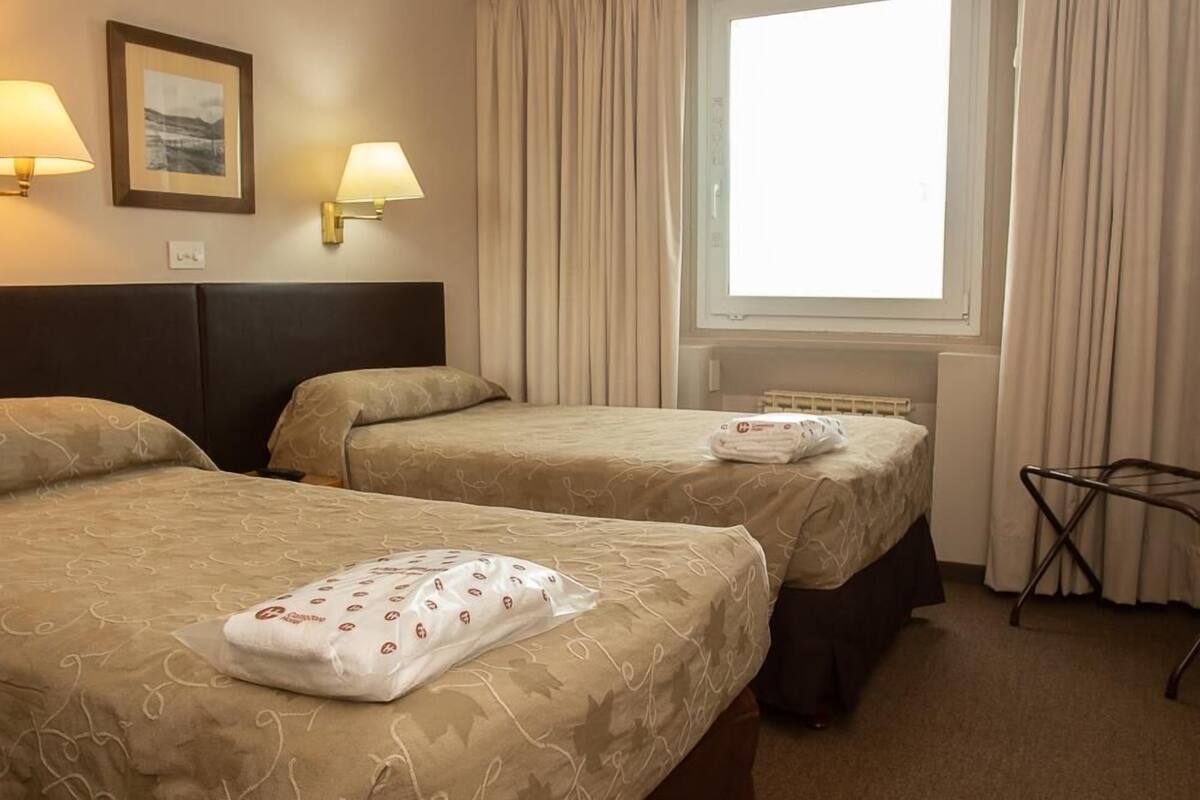 A hotel room with two beds, a window with curtains, wall-mounted lamps, and a framed picture.