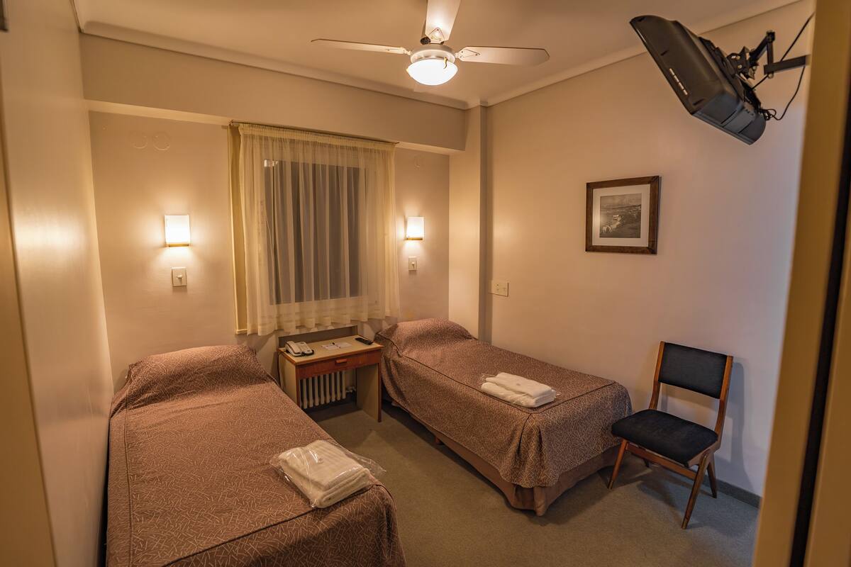 A hotel room with two single beds, a desk with a telephone, a chair, a ceiling fan, and a projector mounted on the wall.