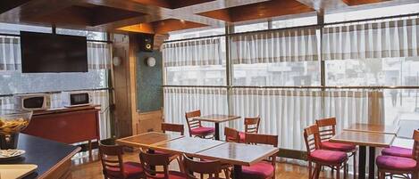 Un comedor con mesas y sillas de madera, una cocina con microondas y ventanas amplias con cortinas.