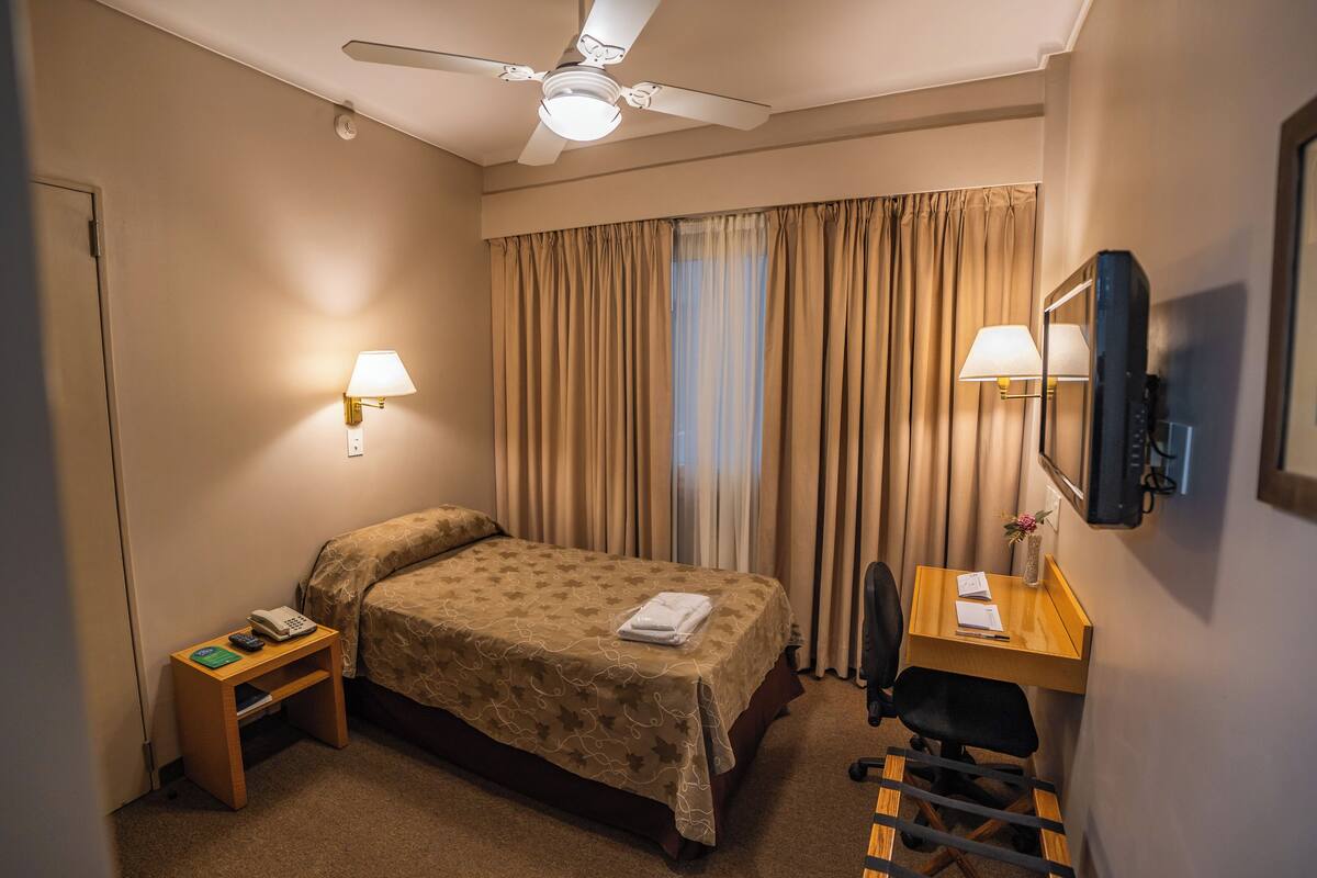 A hotel room with a bed, desk, chair, ceiling fan, and two wall-mounted lamps.