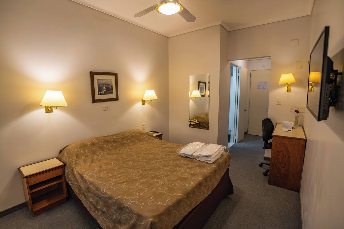 A hotel room with a bed, bedside lamps, a desk, a chair, a mirror, and a ceiling fan.