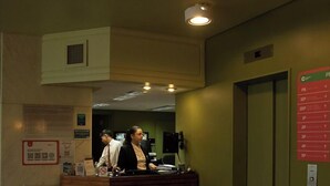 A reception area with a wooden counter, two staff members, a trash bin, and an elevator with a red sign indicating floor levels. - Comodoro Hotel (Comodoro Rivadavia)
