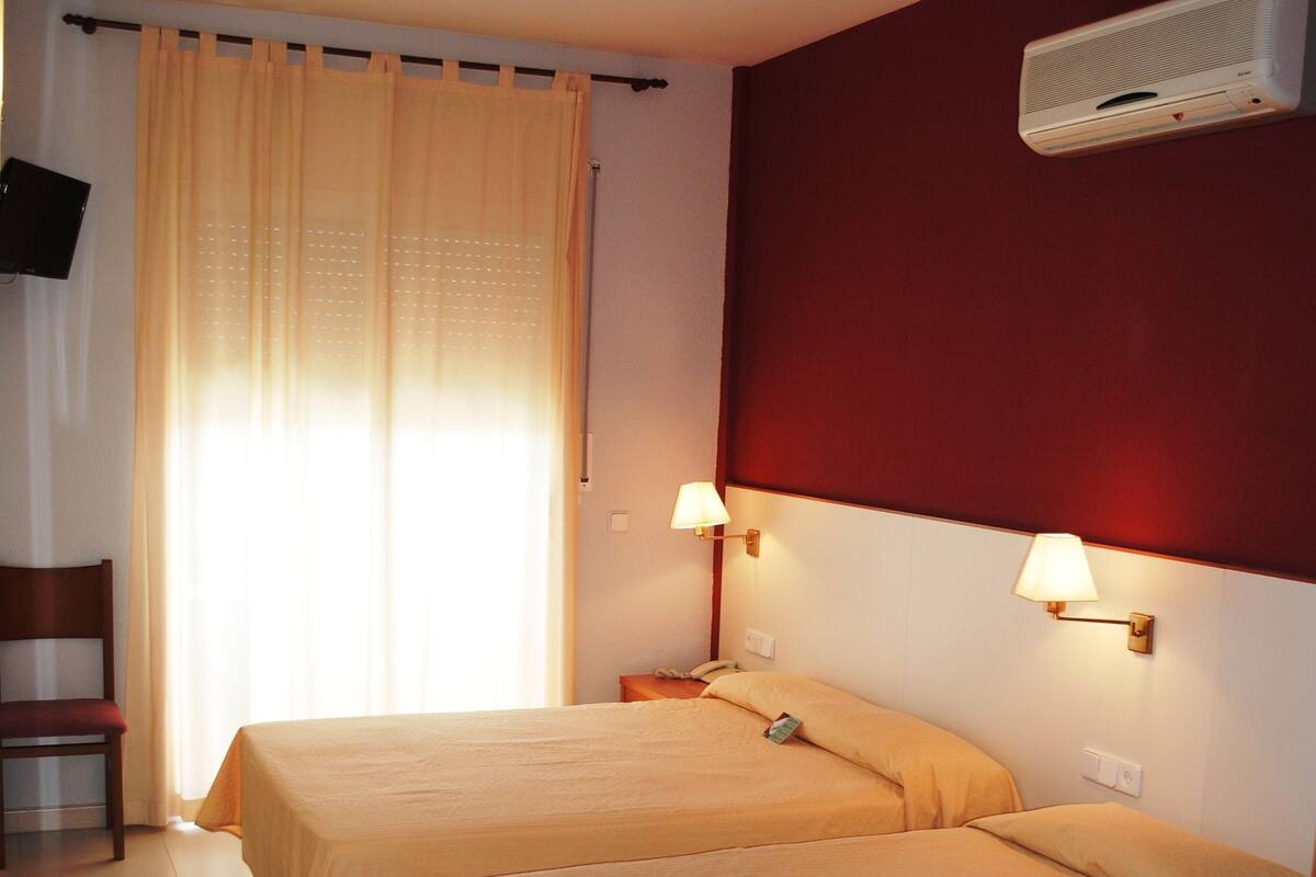 A hotel room with two beds, a TV, a chair, a window with curtains, and an air conditioning unit.