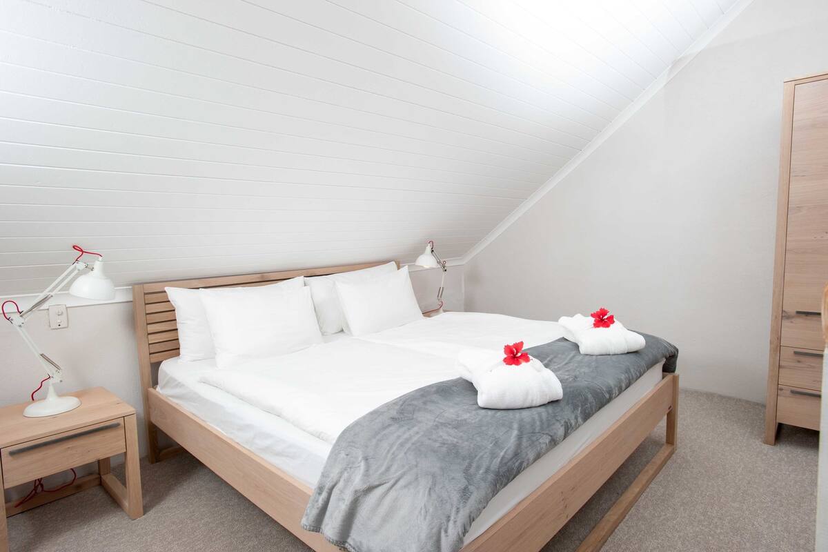 A neatly made bed with white bedding and a gray throw, a wooden nightstand with a lamp, and a wooden dresser in a room with white walls and sloped ceiling.