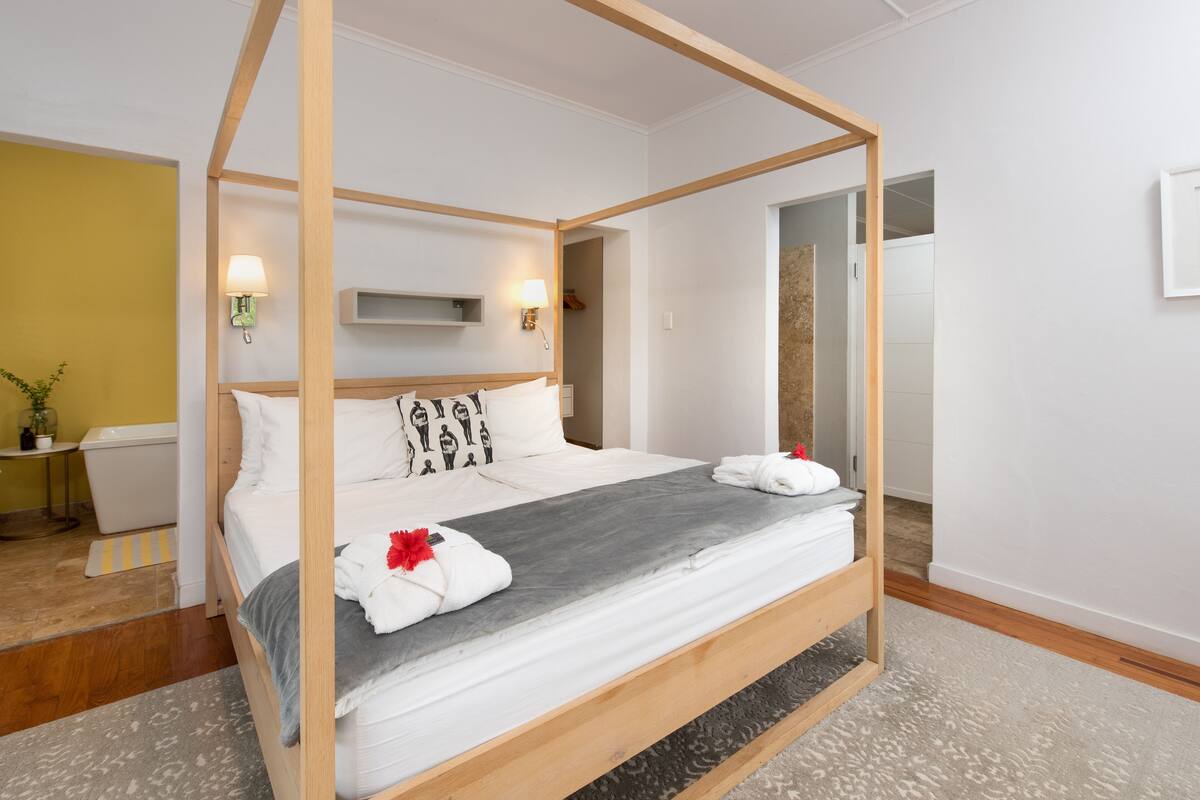 A four-poster bed with white linens and a grey runner, a bedside table with a lamp, and a view of a bathroom and hallway.