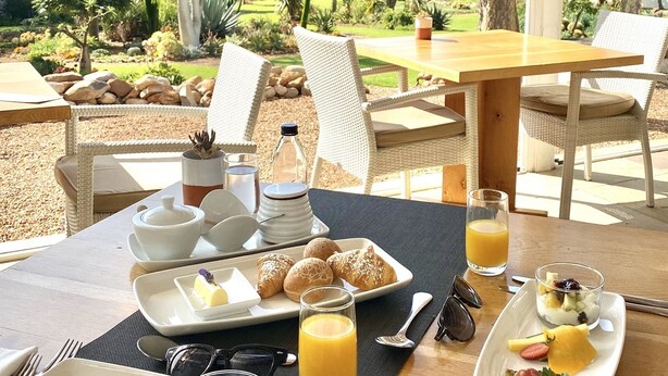 Un desayuno con cruasanes, fruta y jugo sobre una mesa con vistas a un jardín y montañas.