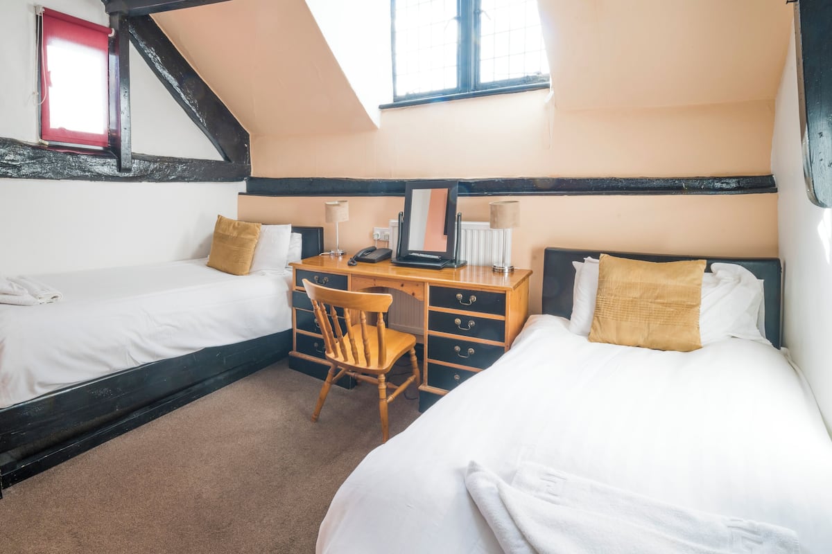 Une chambre avec deux lits, un bureau et une chaise. Il y a une fenêtre avec un rideau rouge et une lucarne.