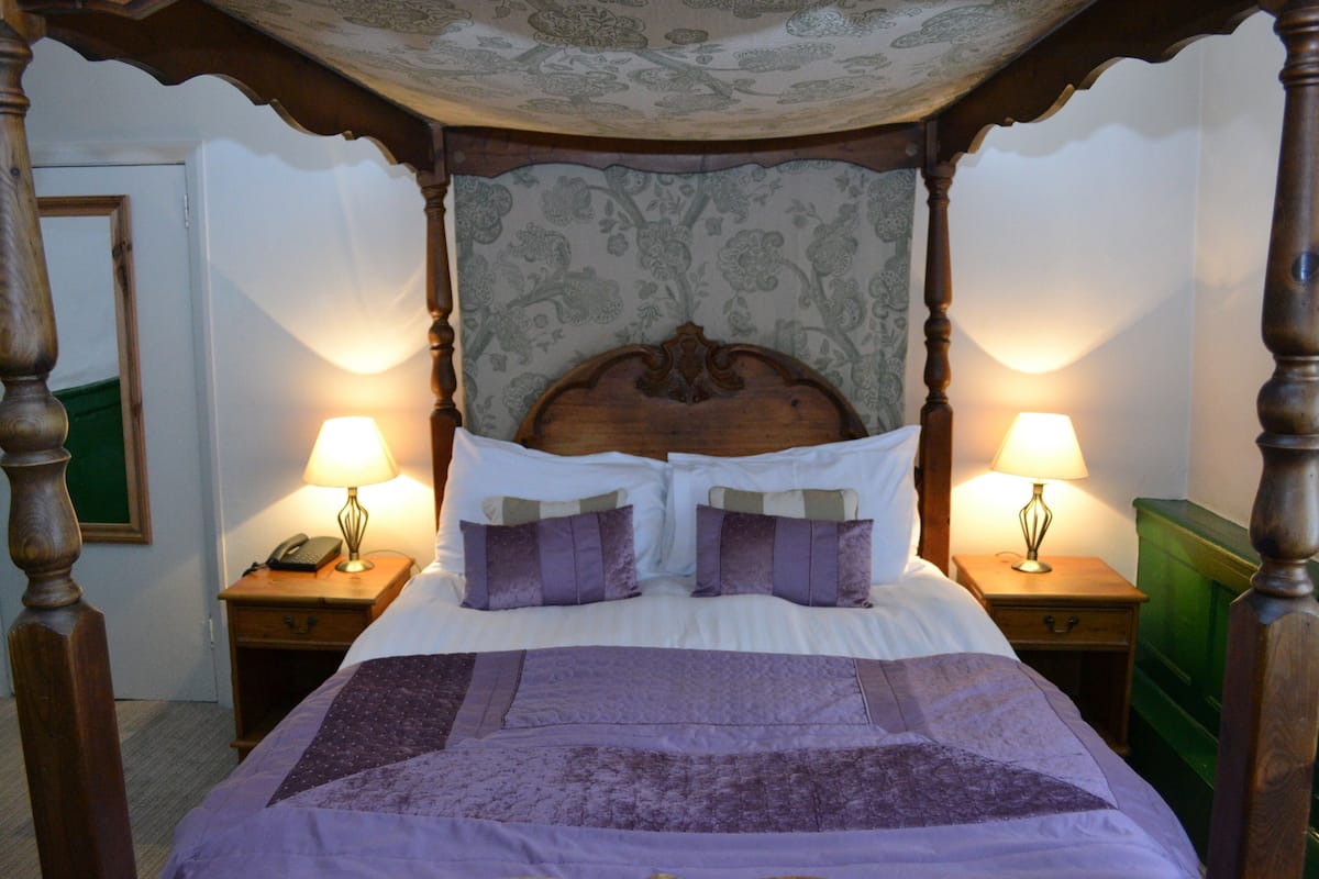 Un lit à baldaquin avec une tête de lit à motifs floraux, deux tables de chevet avec des lampes et une commode verte.