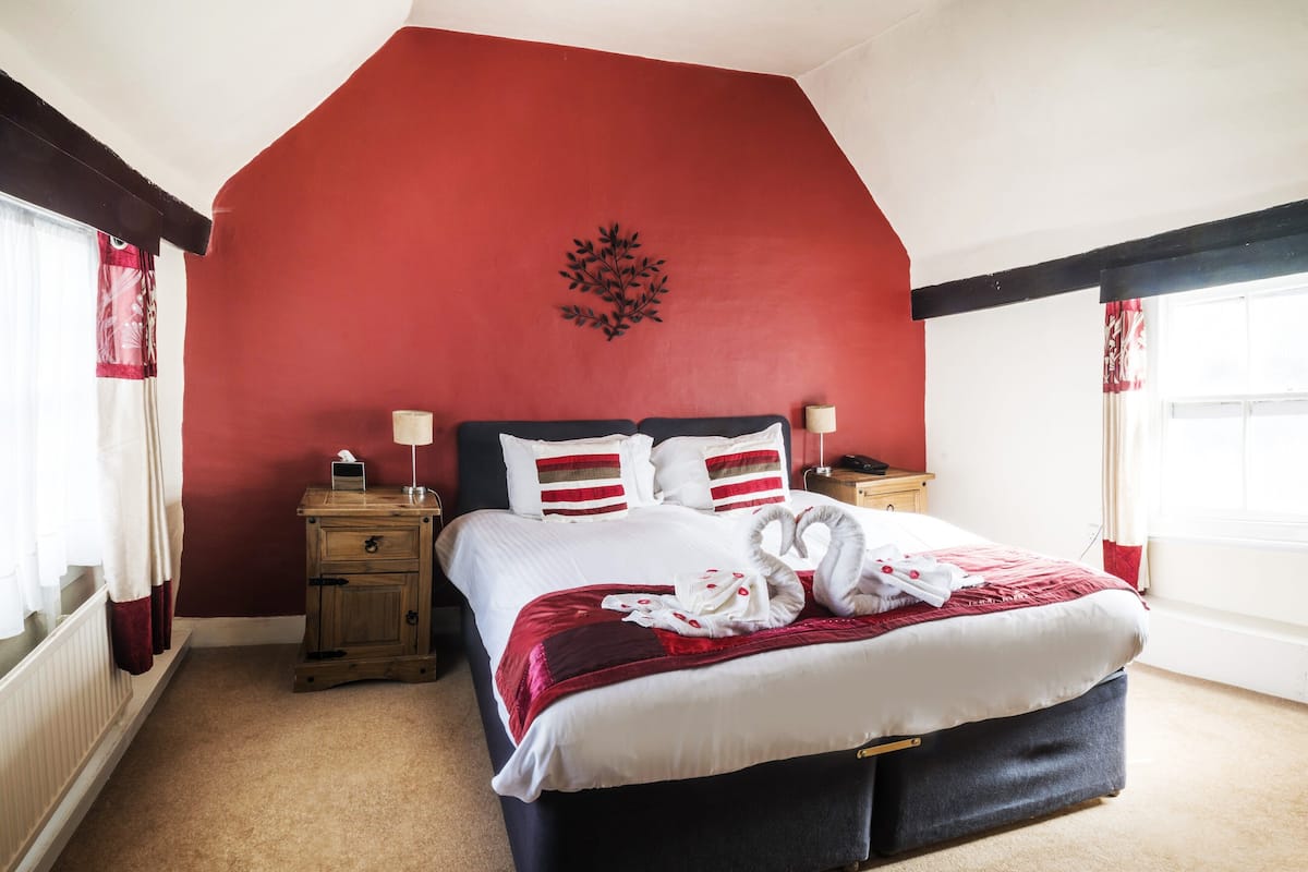 Une chambre avec un mur d’accent rouge, un lit avec une literie blanche, deux tables de chevet avec des lampes et une fenêtre avec des rideaux.