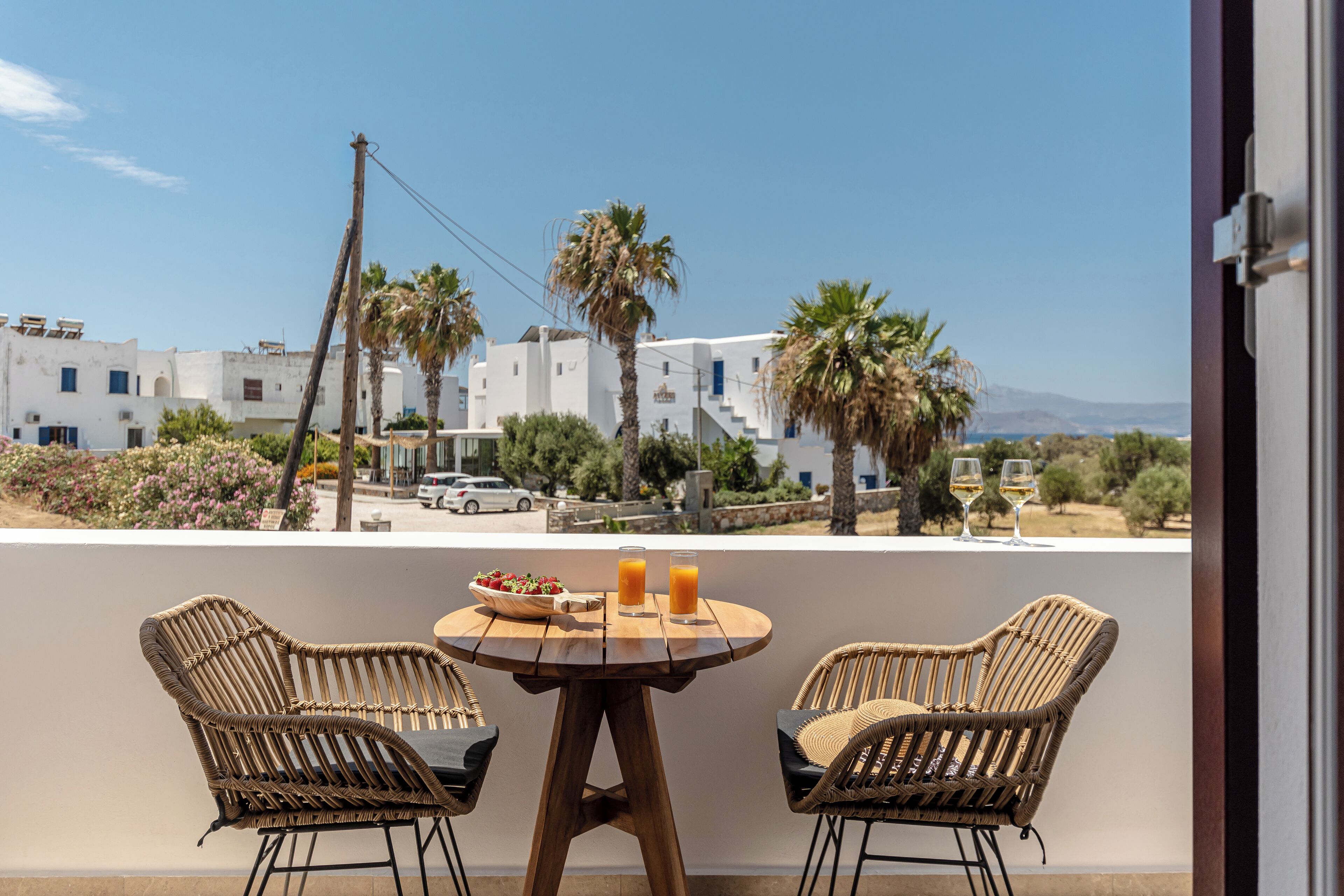 A balcony with wicker chairs and a table, overlooking a coastal town with white buildings and palm trees.
