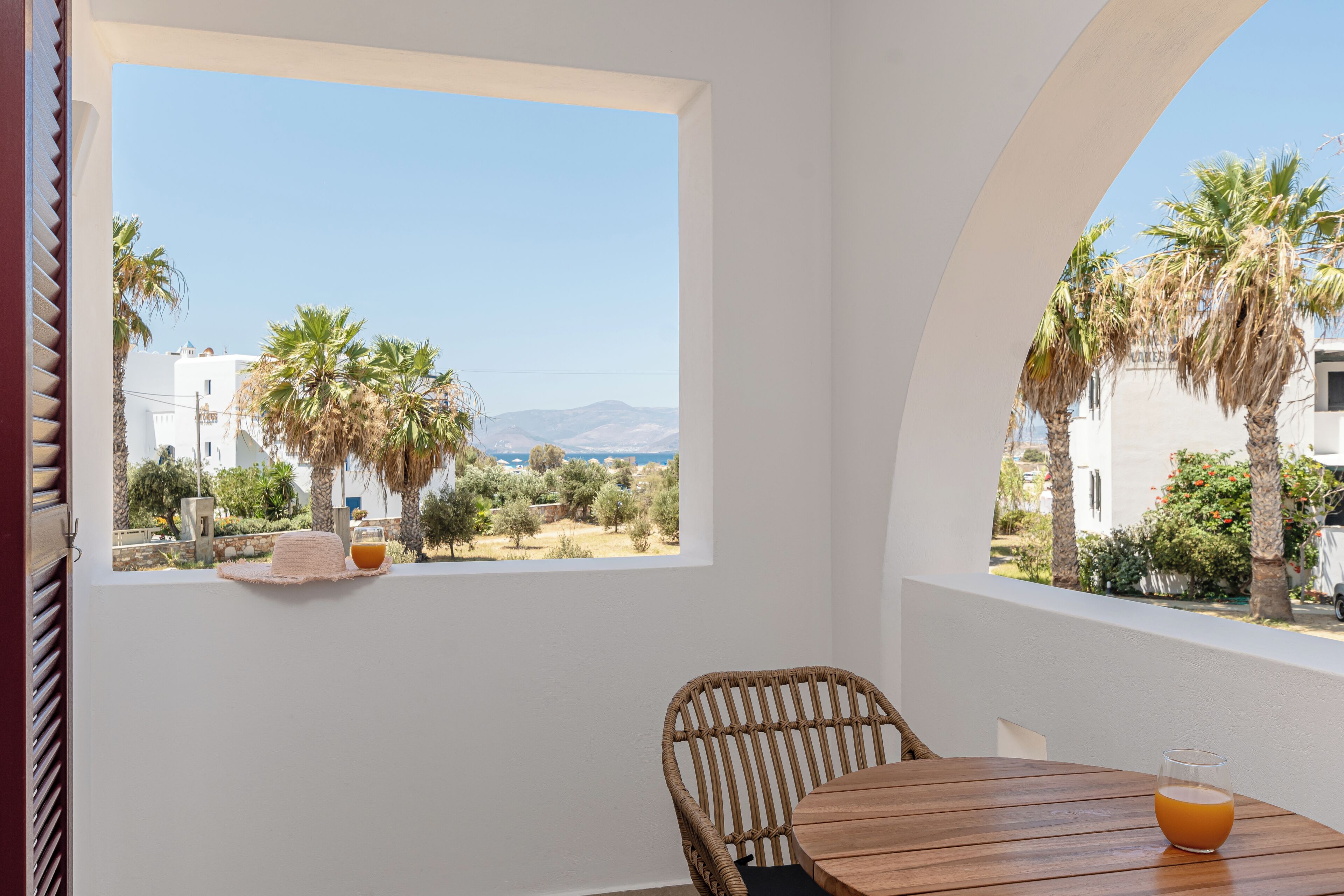 A room with a view of palm trees and buildings, a wicker chair, a wooden table, and a glass of orange juice.