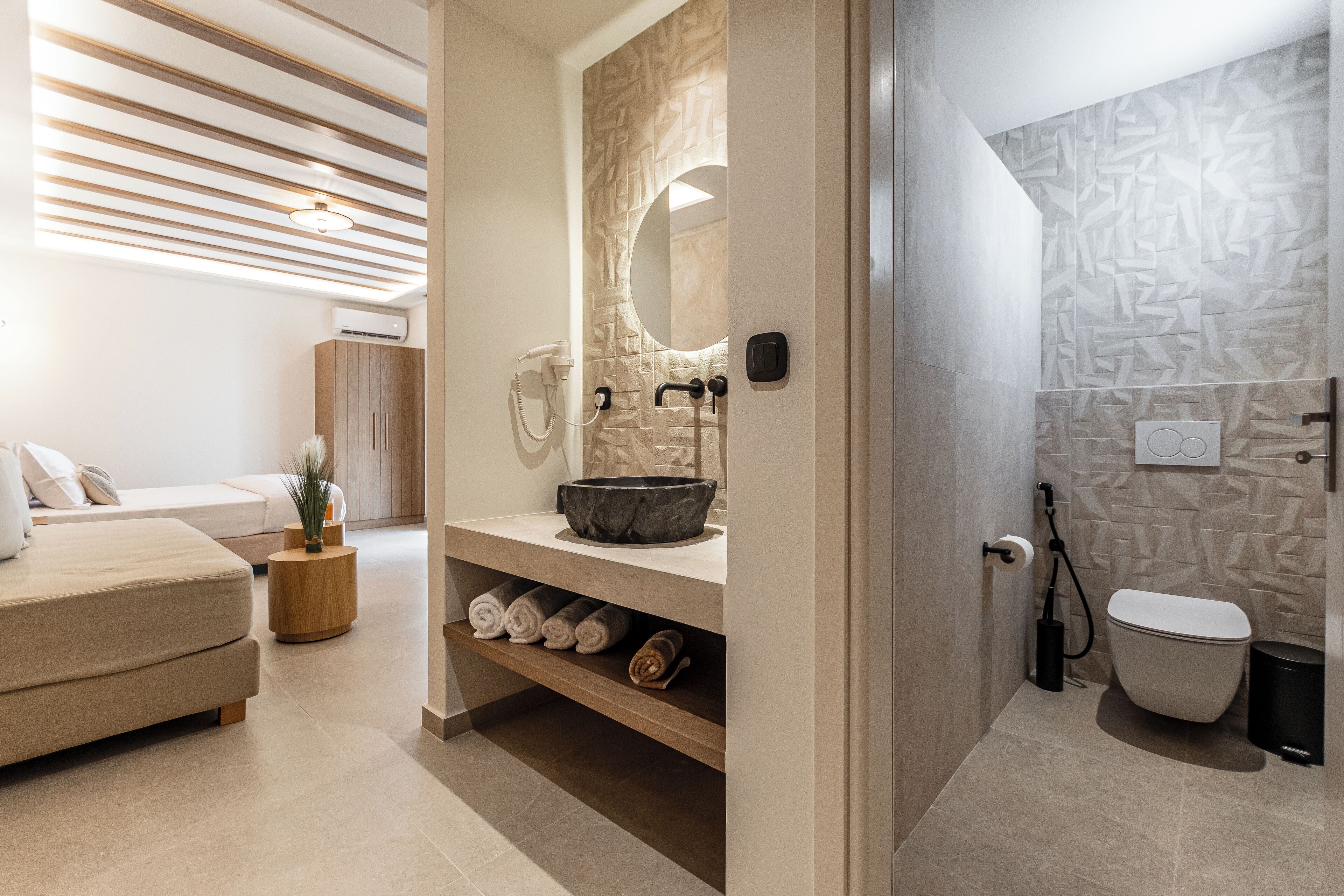Un baño moderno con un espejo grande, un lavamanos y un inodoro. Se aprecia un perchero con toallas y un taburete de madera.