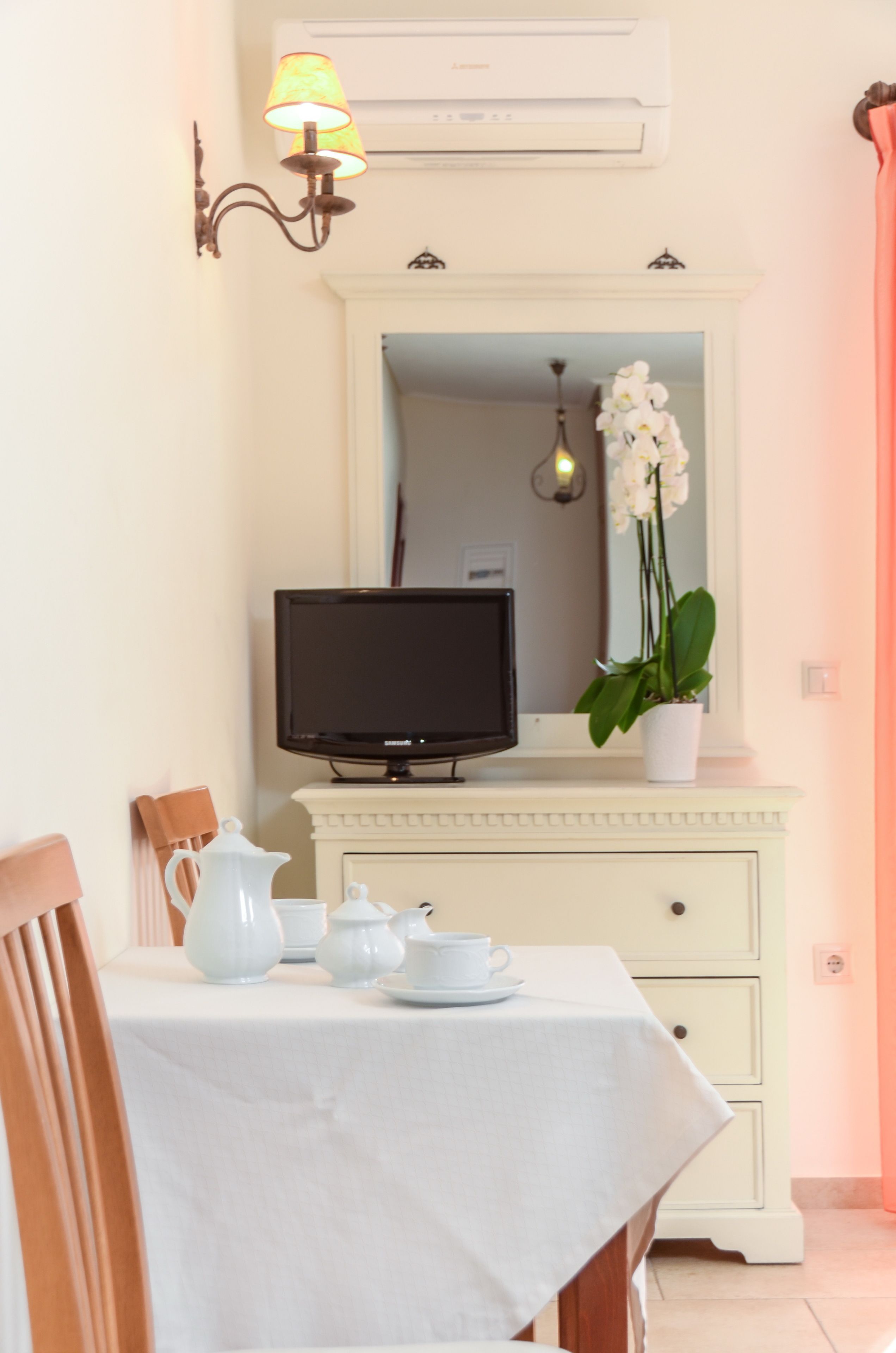 Un comedor con una mesa blanca preparada para el té, un televisor sobre un mueble y una orquídea en maceta.