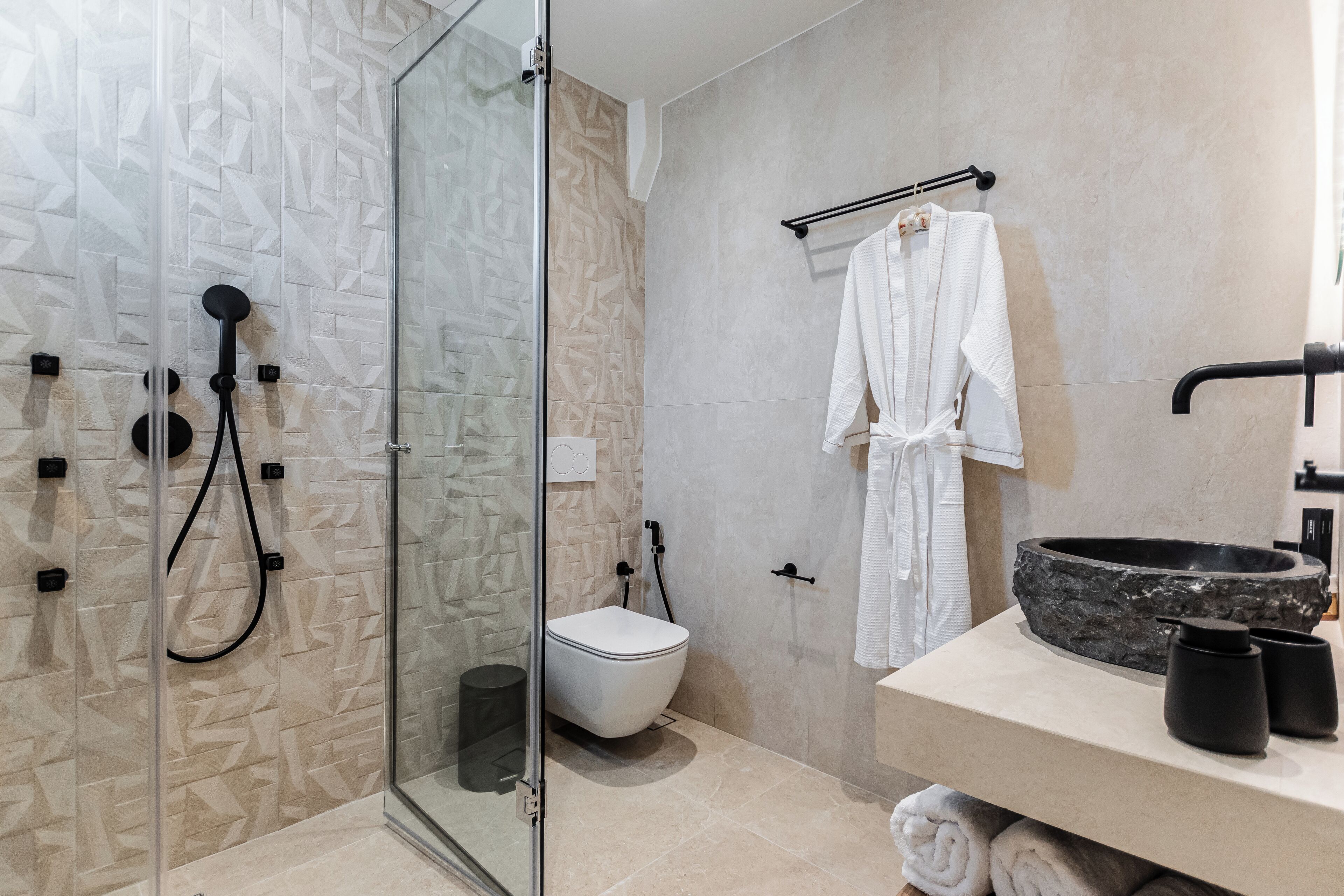 A modern bathroom with a glass-enclosed shower, a white toilet, and a vanity with a stone basin and black cups.